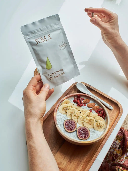 Hand holding a Soma Lion's Mane 60 capsules pouch above a breakfast bowl with figs and banana