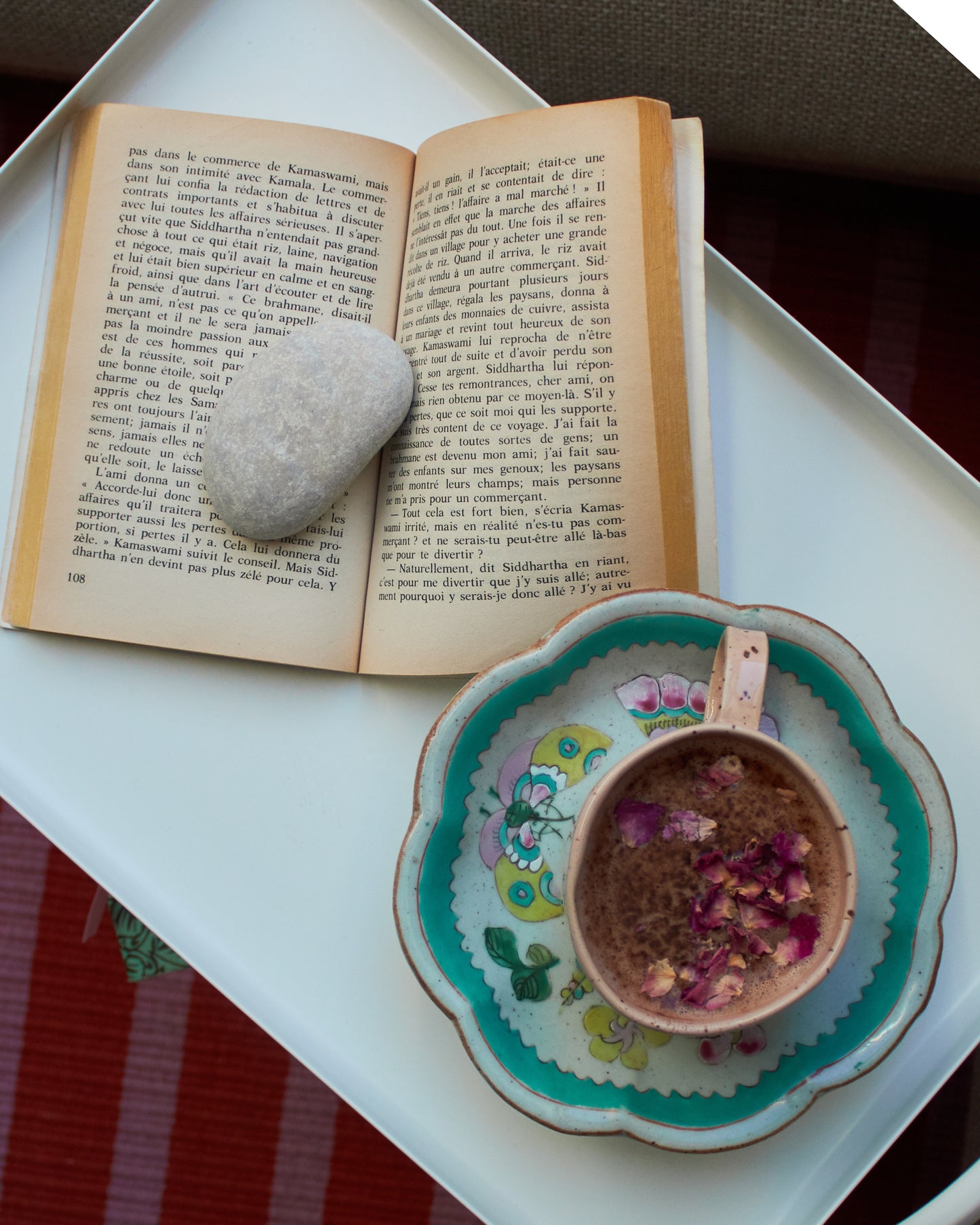 Open book with a stone on top, next to a decorative cup with flowers on a white tray.