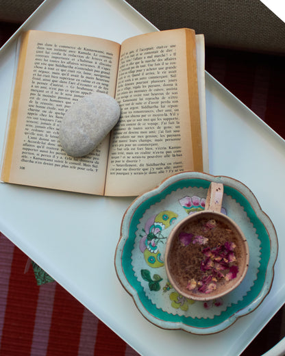 Open book with a stone on top, next to a decorative cup with flowers on a white tray.