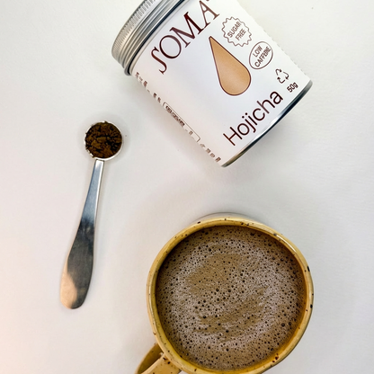 Soma Hojicha powder tin with a spoonful of powder next to a mug of hot beverage on a white background