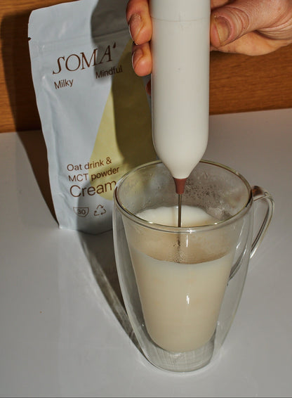 Person using a white handheld device to add a liquid to a glass of milk, with a SOMA Milky Oat drink and MCT powder cream packet in the background.